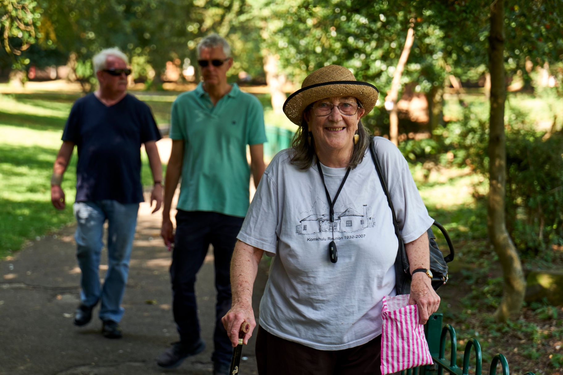 An older woman smiling on a park walk with companions behind her.